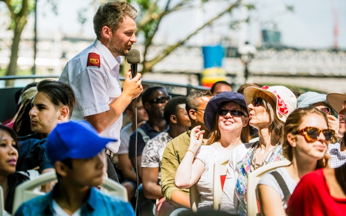 Tour guide speaking to passengers on an open-top bus tour in a city setting.