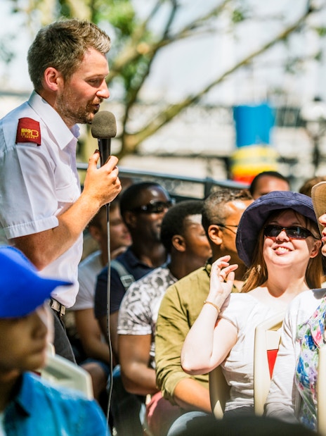 Tour guide speaking to passengers on an open-top bus tour in a city setting.
