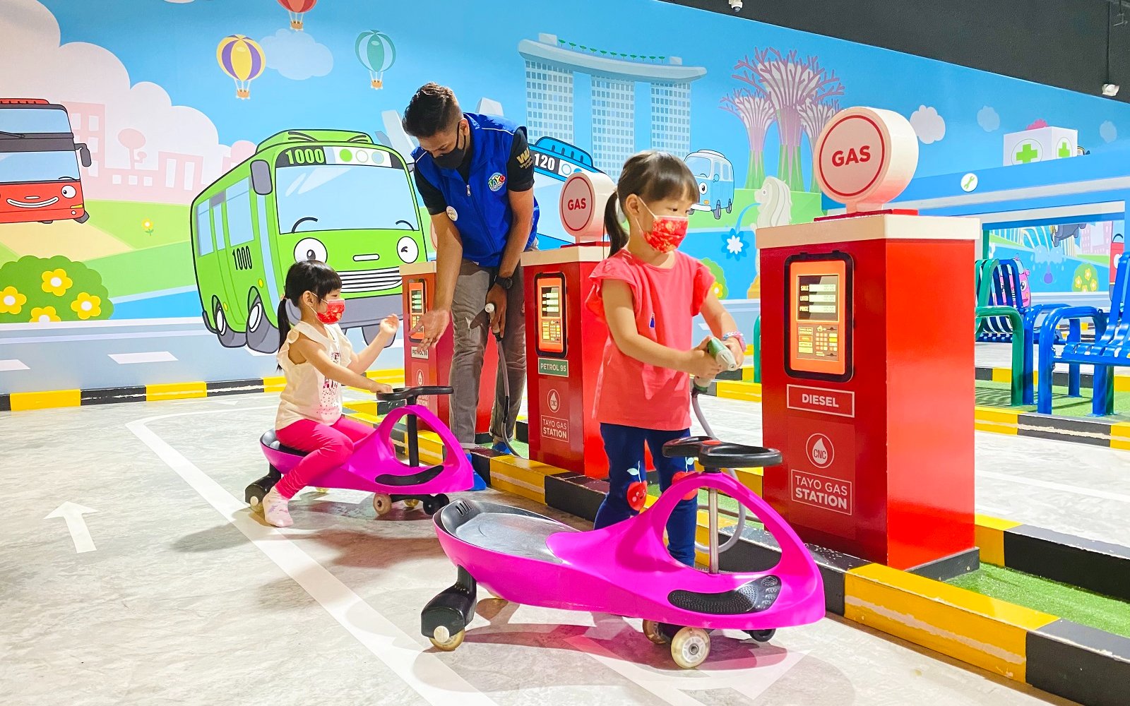 Kids riding toy cycles at Tayo Station gas pump with colorful mural backdrop.