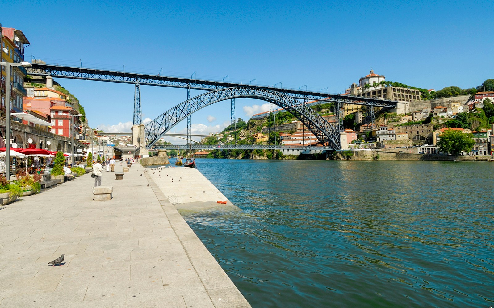 View of Dom Luis I Bridge from Cais da Ribeira, Porto, with riverside promenade.