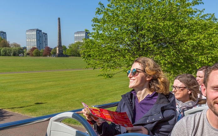 Tourists on an open-top bus in Glasgow, viewing the city's landmarks and greenery.