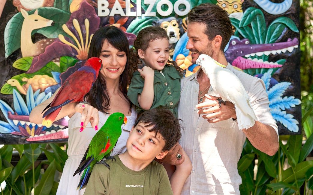 Family with colorful parrots in front of a mural at Bali Zoo.