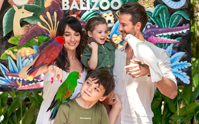 Family with colorful parrots in front of a mural at Bali Zoo.