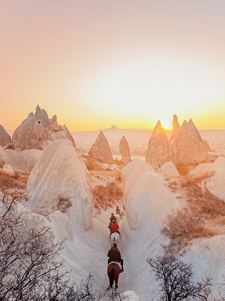 Horseback riders in Cappadocia valley at sunrise with hot air balloons in the sky.