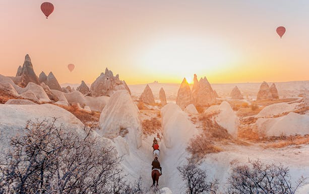 Horseback riders in Cappadocia valley at sunrise with hot air balloons in the sky.