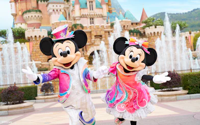 Mickey and Minnie Mouse in colorful outfits at Disneyland Hong Kong with castle backdrop.