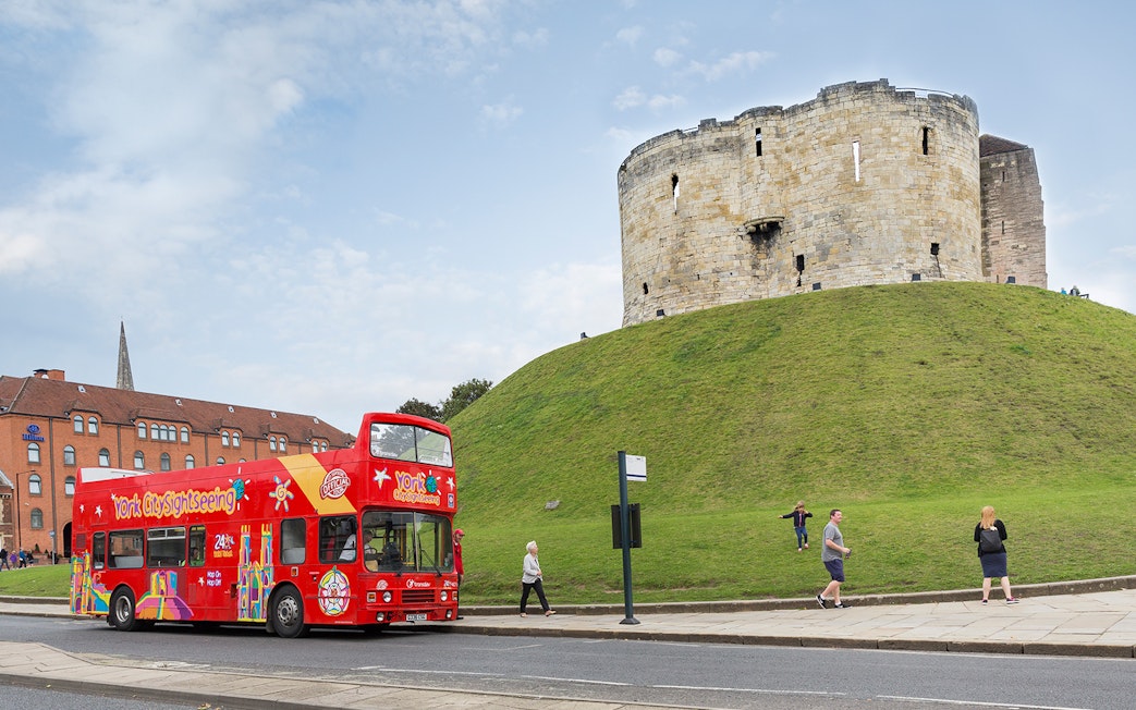 Red double-decker bus on York Hop-On-Hop-Off Tour passing Clifford's Tower in York, England.