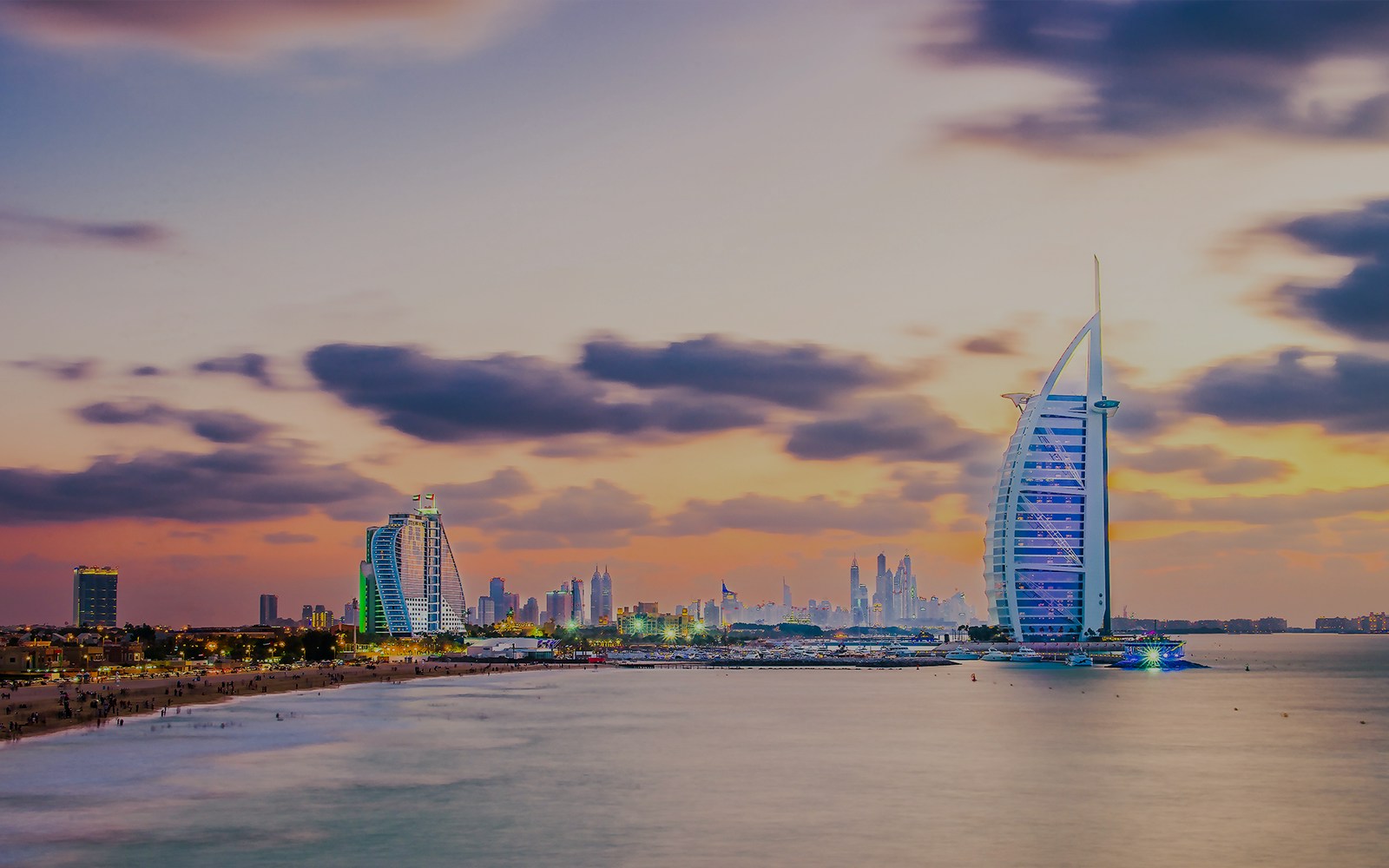 Burj Al Arab illuminated at evening with Dubai skyline in the background.
