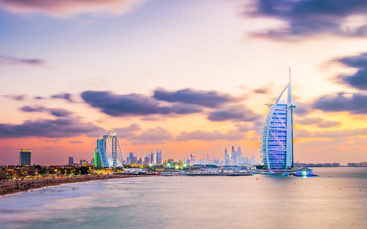 Burj Al Arab illuminated at evening with Dubai skyline in the background.