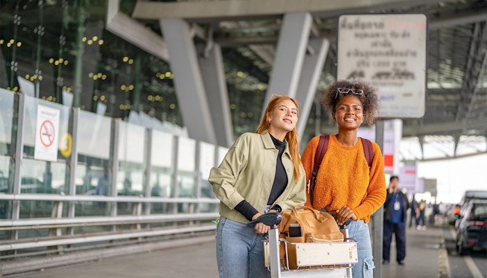 Two women with luggage waiting at airport pickup area.