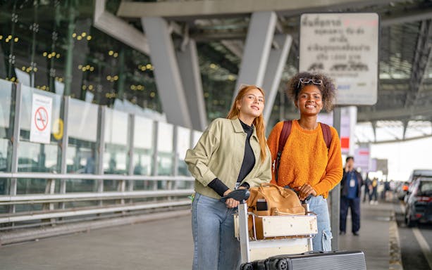 Two women with luggage waiting at airport pickup area.