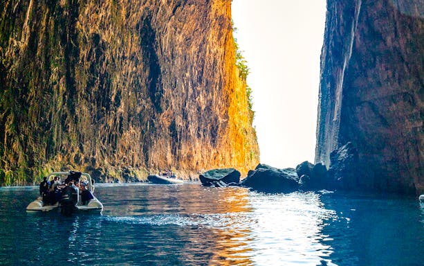 Boat entering Haxhi Ali Caves with towering rock walls and clear blue water.