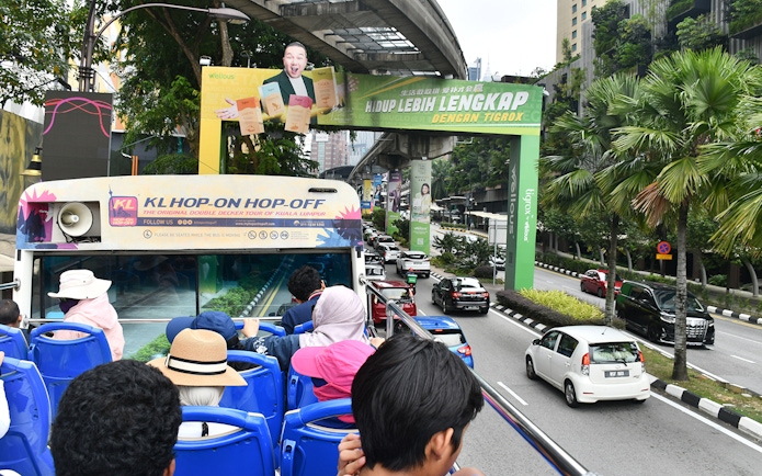 Open-top bus tour in Kuala Lumpur with passengers viewing city streets and greenery.