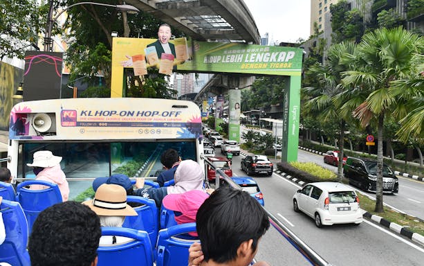 Open-top bus tour in Kuala Lumpur with passengers viewing city streets and greenery.