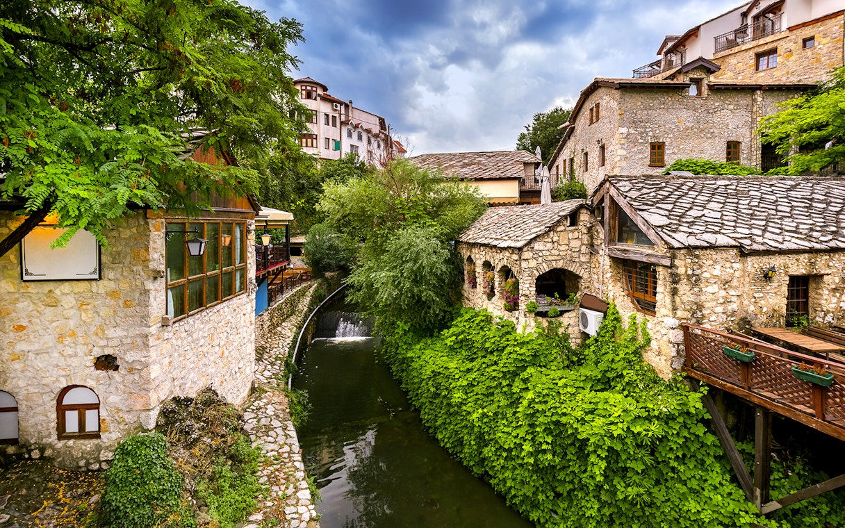 Stone buildings and lush greenery along a river in Mostar, part of the guided tour to Kravica Waterfall.
