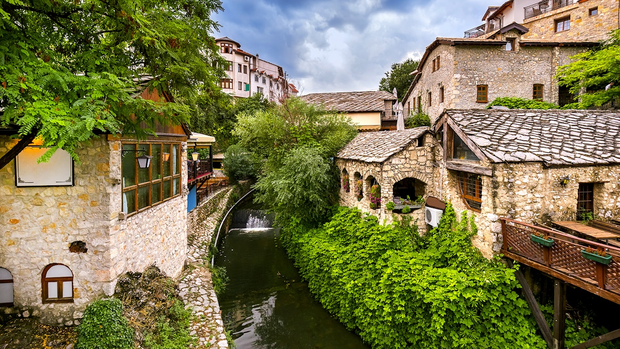 Stone buildings and lush greenery along a river in Mostar, part of the guided tour to Kravica Waterfall.