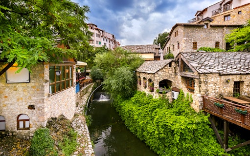 Stone buildings and lush greenery along a river in Mostar, part of the guided tour to Kravica Waterfall.