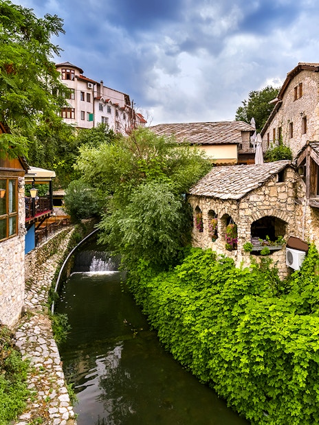Stone buildings and lush greenery along a river in Mostar, part of the guided tour to Kravica Waterfall.