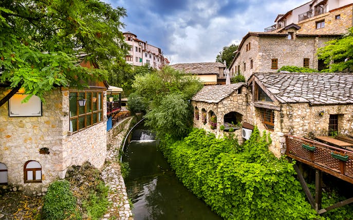 Stone buildings and lush greenery along a river in Mostar, part of the guided tour to Kravica Waterfall.