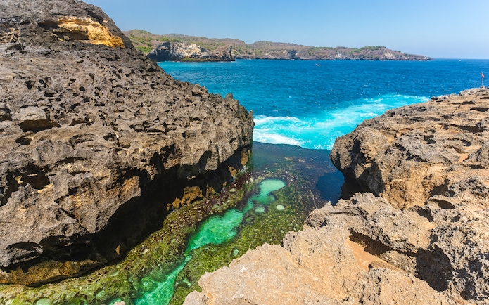 Natural rock pool at Angel's Billabong with clear turquoise water, Bali coastline in background.