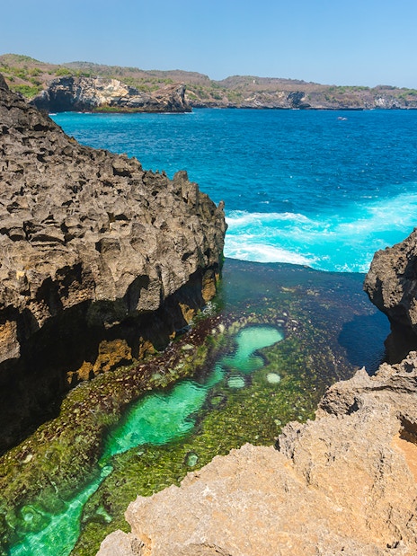 Natural rock pool at Angel's Billabong with clear turquoise water, Bali coastline in background.