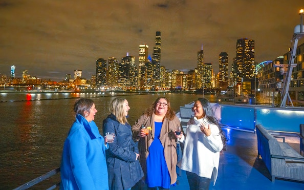 Group enjoying drinks on a Chicago dinner cruise with city skyline at night.