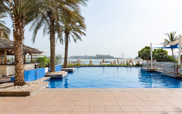 Riva Beach Club pool with palm trees and view of Dubai skyline.