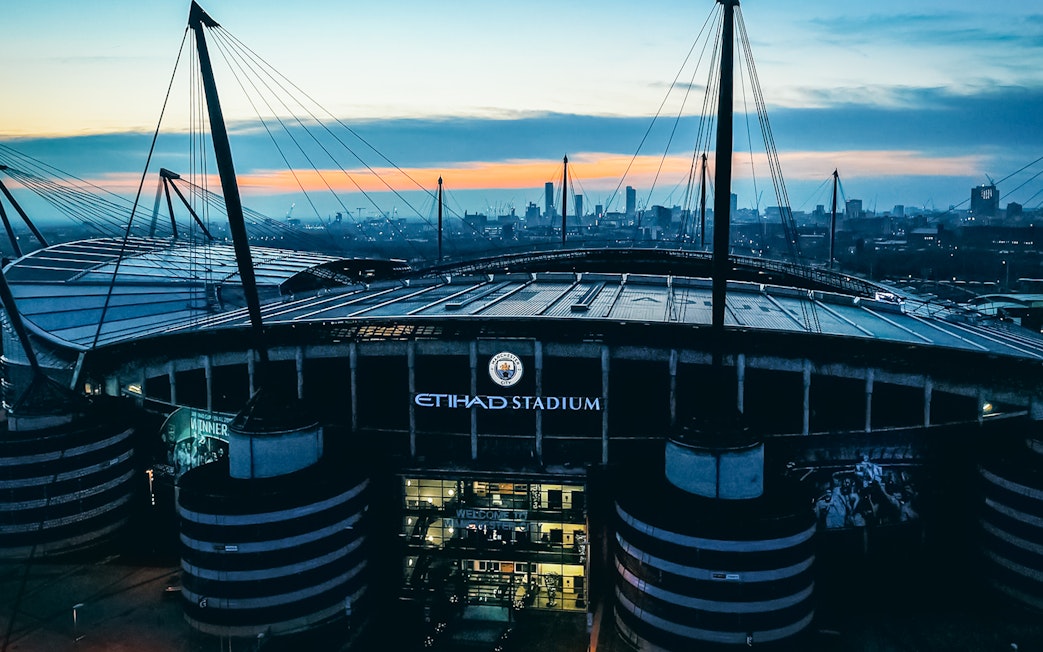 Etihad Stadium in Manchester at sunset with city skyline in background.
