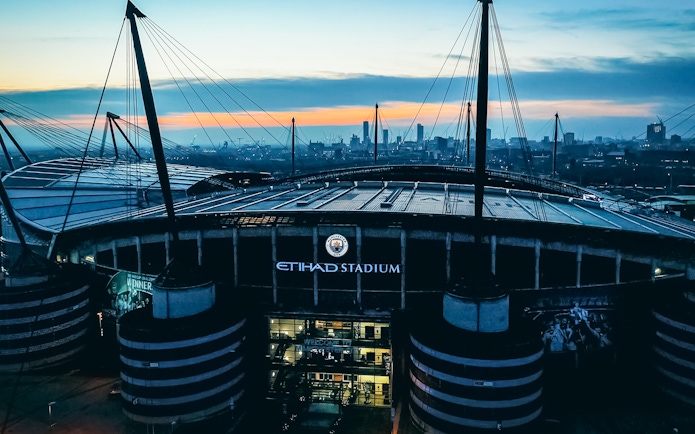 Etihad Stadium in Manchester at sunset with city skyline in background.
