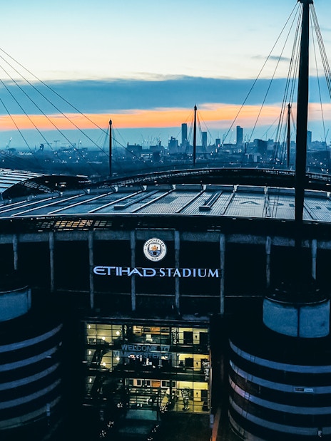 Etihad Stadium in Manchester at sunset with city skyline in background.