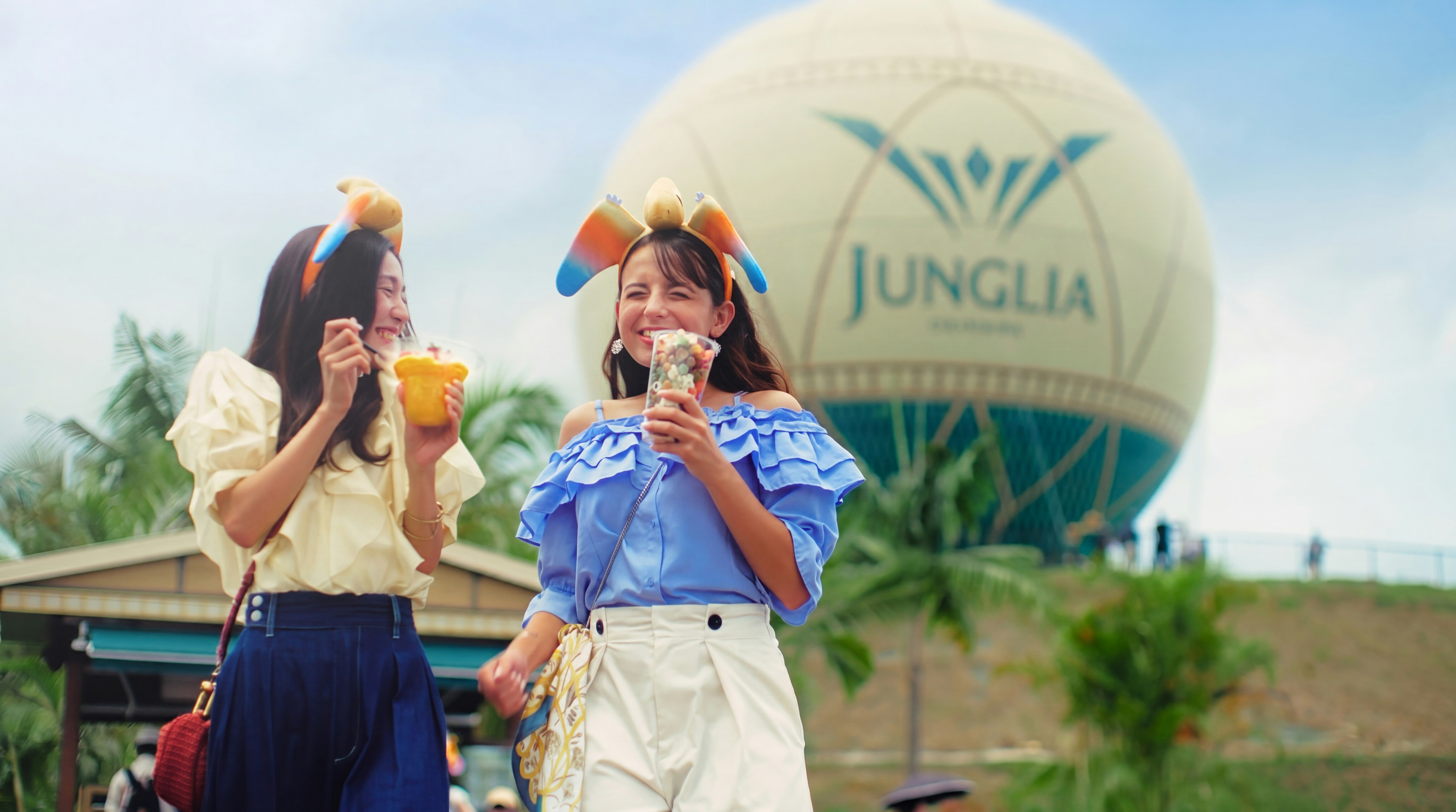 Two visitors enjoying snacks at JUNGLIA OKINAWA Park with the park's balloon in the background.