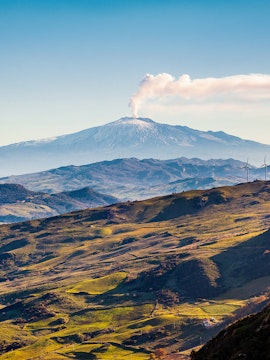 Mount Etna with smoke rising, surrounded by green hills in Sicily, Italy.