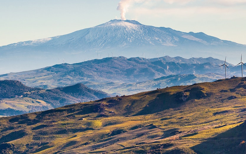 Mount Etna with smoke rising, surrounded by green hills in Sicily, Italy.