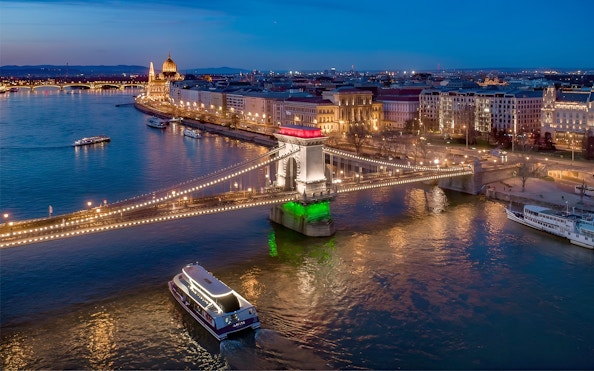 Cruise boat on Danube River near illuminated Chain Bridge in Budapest at dusk.