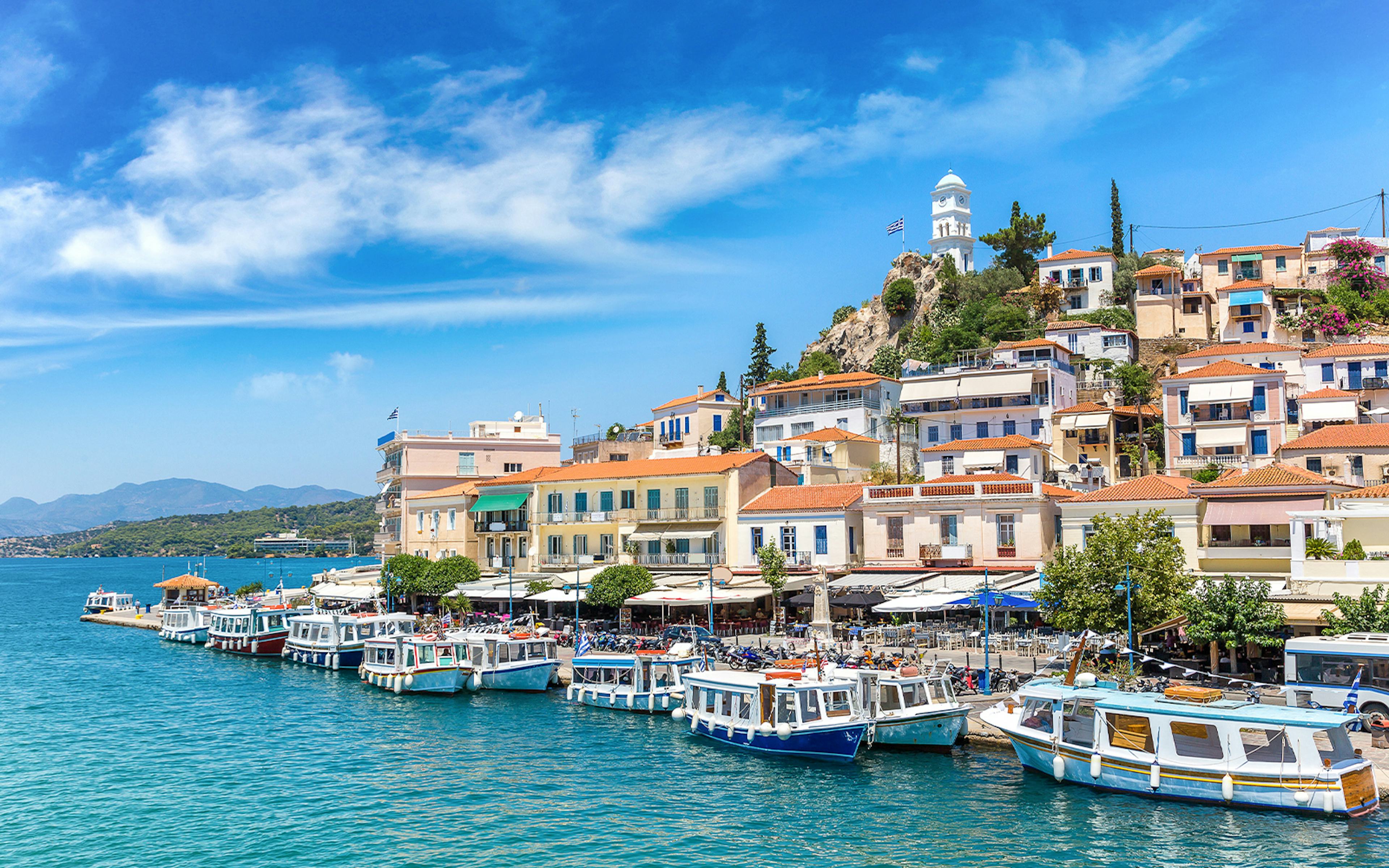 Boats docked along the waterfront in Poros, Greece, with hillside houses and a clock tower.