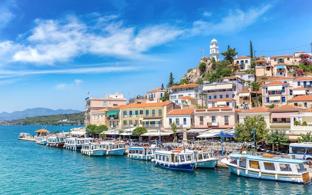Boats docked along the waterfront in Poros, Greece, with hillside houses and a clock tower.