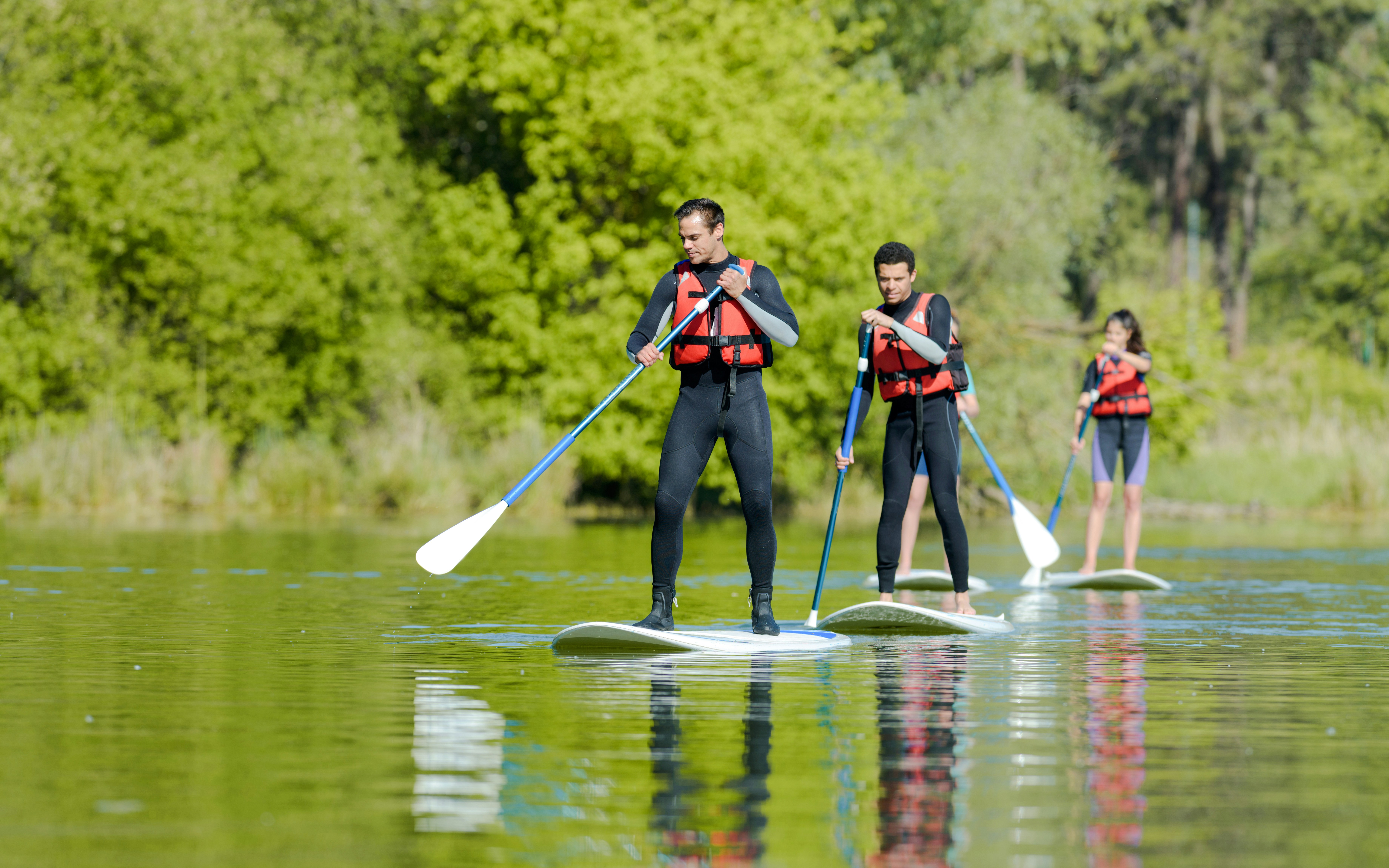 People paddle boarding on a calm lake wearing life jackets, surrounded by lush greenery.
