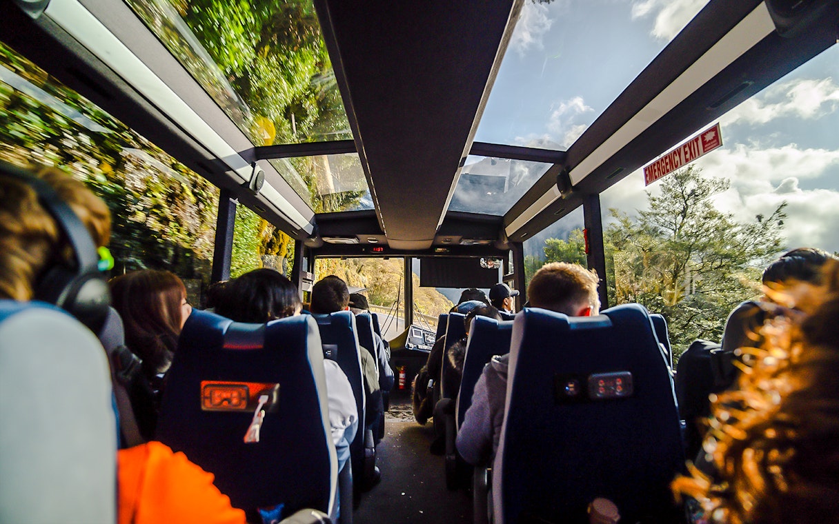 Passengers on a glass-roof bus traveling through Milford Sound scenery.
