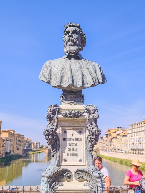 Bust of Benvenuto Cellini on Ponte Vecchio, Florence, with tourists and river view.