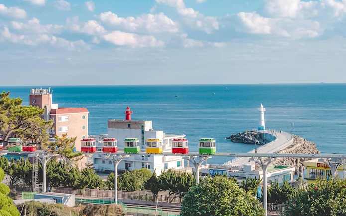 Blueline Park Sky Capsule overlooking Busan coastline with lighthouse and colorful buildings.
