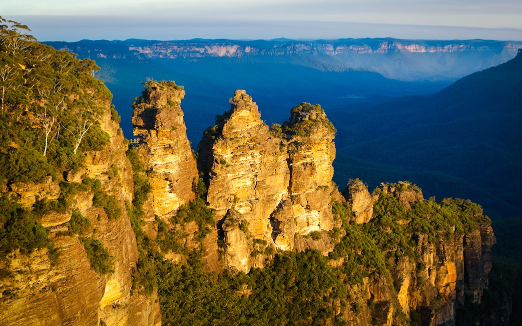 Three Sisters rock formation in Blue Mountains, Australia, with forested valley backdrop.