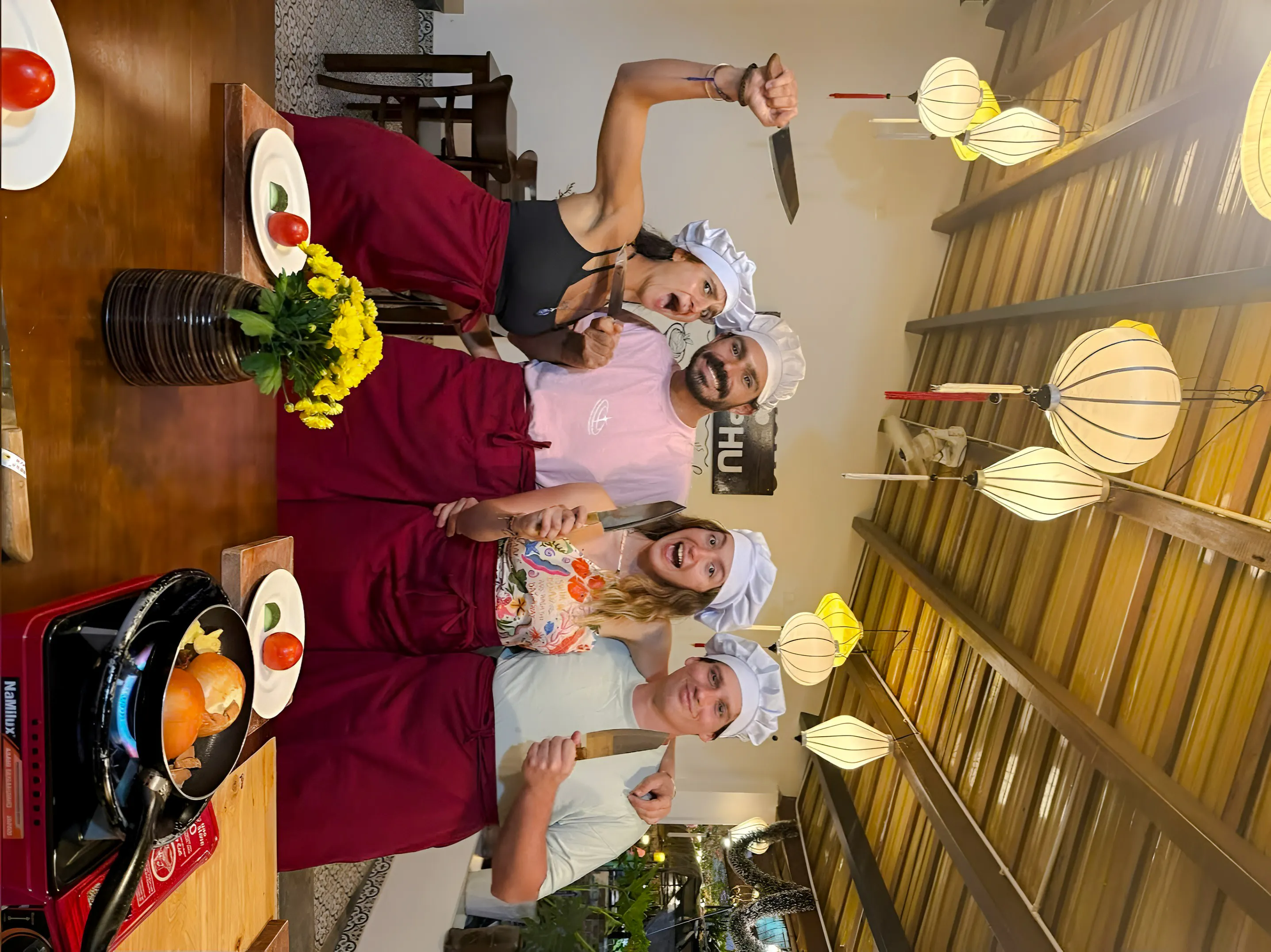 Group cooking class participants in Hoi An wearing chef hats and aprons, holding knives.