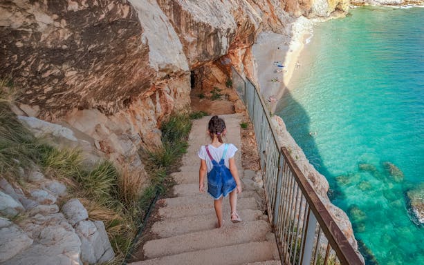 Child walking down stairs to Pasjača Beach, Croatia, with rocky cliffs and turquoise water.