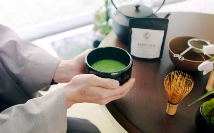 Hands holding a bowl of matcha tea with a whisk and tea set on a table in Tokyo.