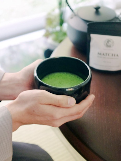 Hands holding a bowl of matcha tea with a whisk and tea set on a table in Tokyo.