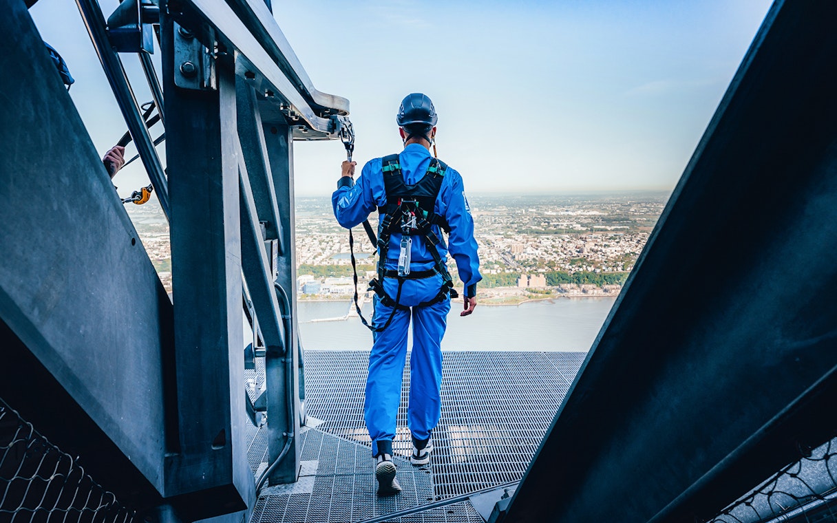 Person in safety gear walking on a high platform overlooking a city and river.