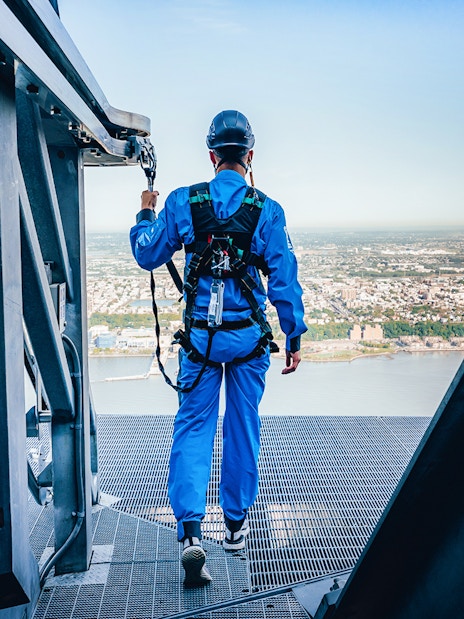 Person in safety gear walking on a high platform overlooking a city and river.