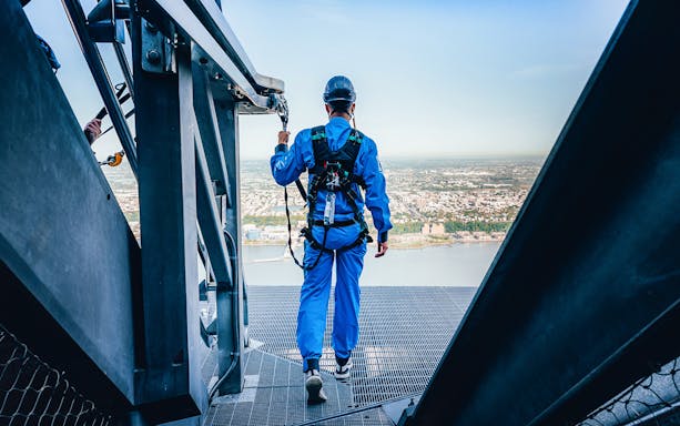 Person in safety gear walking on a high platform overlooking a city and river.