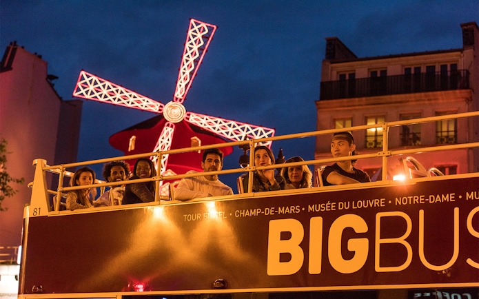 Open-top bus tour passing Moulin Rouge at night in Paris.