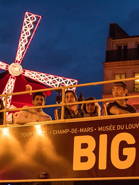 Open-top bus tour passing Moulin Rouge at night in Paris.